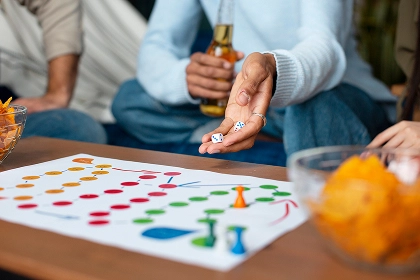 Een groep vrienden speelt een kleurrijk bordspel. De een houdt dobbelstenen vast, terwijl een ander iets te drinken heeft. De sfeer is ontspannen en gezellig, met wat hapjes binnen handbereik.